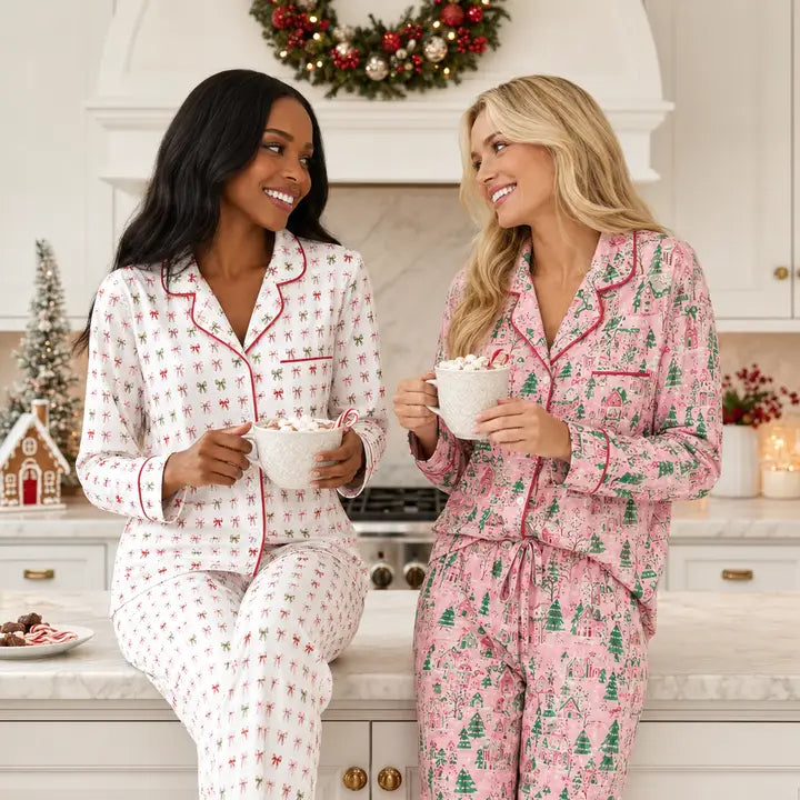 Two women wearing matching holiday-themed pajama sets in a bright kitchen. One set is white with red trim and small red motifs, and the other is pink with red trim and green tree motifs.